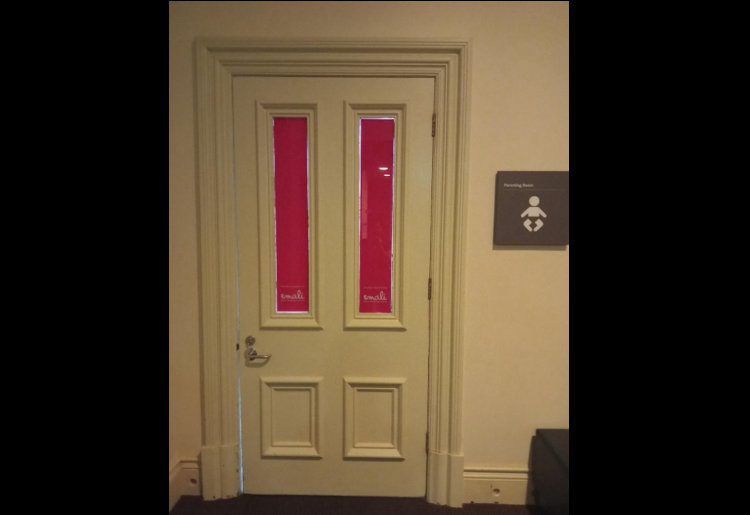 External view of the Parents Room. The door is a light cream colour with two vertical rectangular windows covered by pink panels. To the right of the door, a grey sign with a white icon indicates a parenting room.